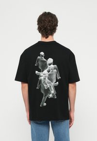 Μαύρο μπλουζάκι με γραφικό σχέδιο πολλών φιγούρων σε κίνηση στην πλάτη. Κοντά μανίκια, στρογγυλή λαιμόκοψη, κατασκευασμένο από βαμβάκι.
