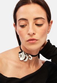 Sort choker med skinnende metallisk blomsterpendant, der har buede, overlappende kronblade. Modellen bærer en off-shoulder sort top.