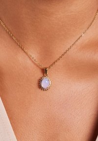 Gouden ketting met een ovale, lichtblauwe stenen hanger omringd door kleine heldere stenen, rustend op blote huid. Ketting met ronde schakels.