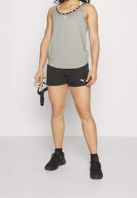 Grå atletisk tanktop med svarta detaljer, svarta shorts och svarta sneakers. Modellen håller i träningsband och visar upp en sportig outfit.