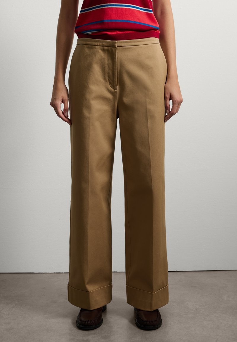 Personne portant un pantalon kaki taille haute à jambes larges avec revers retroussés, associé à des chaussures marron foncé et un haut rayé rouge, bleu et blanc.