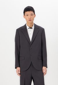 Donkergrijze op maat gemaakte blazer met een gestructureerd ontwerp, revers met notches, twee voorzakken en twee knopen. Gelaatst met een wit shirt.