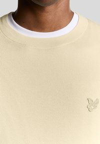 Sweatshirt bege em malha com gola redonda, colarinho branco sobreposto e pequeno logótipo bordado no peito esquerdo. Textura suave.