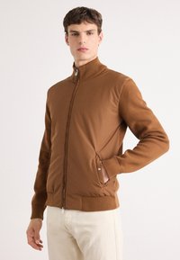 Brūns zip-up jakas ar augstu apkakli, ribotām piedurknēm un sānu kabatām. Gluda tekstūra, pieguļoša forma un delikāts zīmola elements uz piedurknes.