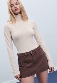 Beige Rollkragenpullover mit langen Ärmeln, kombiniert mit einem braunen Jeansrock. Der Rock hat vordere Taschen und einen Knopfverschluss.