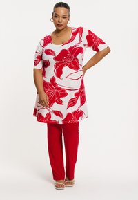 Weiße Tunika mit rotem Blumenmuster, kurzen Ärmeln und runder Ausschnitt, kombiniert mit roten Hosen. Offene weiße Sandalen runden das Outfit ab.