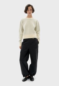 Une personne se tient debout, les mains dans les poches, portant un pull à col rond crème, un pantalon cargo noir ample avec des ourlets à cordon, et des chaussures noires à enfiler.