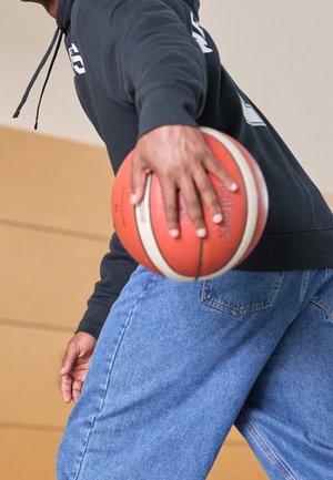 Homme en sweat à capuche noir et jean bleu tenant un ballon de basket orange, prêt à dribbler ou à passer en intérieur.