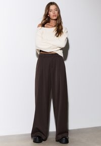 Pull en maille oversized couleur blanc cassé avec encolure large. Pantalon plissé taille haute couleur marron foncé. Chaussures à plateforme épaisses noires.