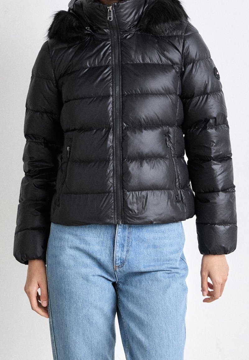 Veste matelassée noire avec capuche bordée de fausse fourrure, portée avec un jean bleu clair, fermeture éclair devant et poches zippées.