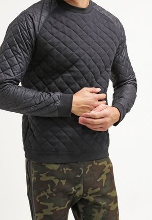 Man draagt een zwart doorgestikt sweatshirt en groene camouflagebroek en houdt zijn buik met beide handen vast.