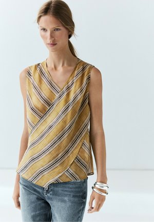 Donna che indossa un top senza maniche a righe gialle e marroni con jeans blu e grandi braccialetti d'argento al polso destro.