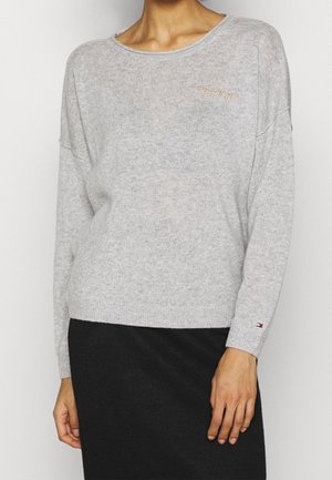 Personne portant un pull léger à manches longues gris clair avec de petits logos brodés sur la poitrine et la manche, associé à une jupe noire.