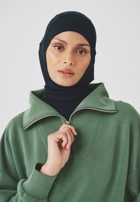 Green sweatshirt with a high collar and half-zip, worn over a black fitted hijab. Smooth texture and relaxed fit, with thumbholes in sleeves.