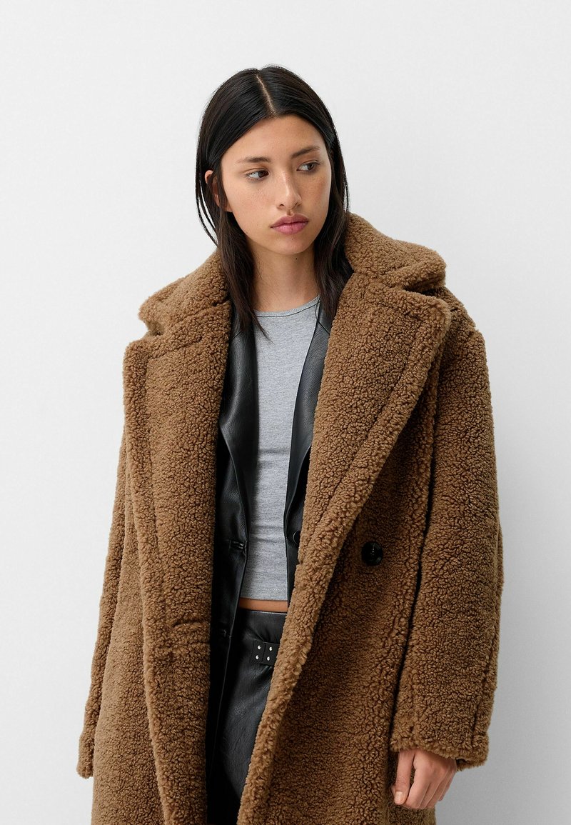 Manteau en teddy marron avec un extérieur doux et texturé, de grands revers et une coupe décontractée. Présente de gros boutons et est porté sur un haut gris et un pantalon noir.