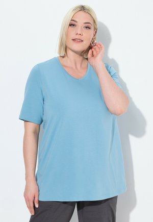 Světle modré tričko s krátkým rukávem a kulatým výstřihem, s volným střihem, vyrobené z měkké látky; nošeno s šedými kalhotami.