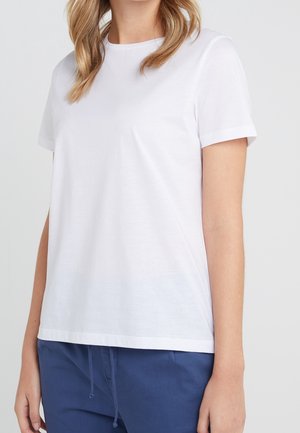 Kobieta ubrana w prosty biały T-shirt z krótkim rękawem oraz granatowe spodnie ze ściągaczem, widoczna od ramion do bioder na jasnym tle.