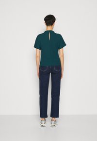 Tyrkysový top s krátkými rukávy a detailem ve tvaru klíčové dírky na zádech, kombinovaný s tmavě modrými džíny a stříbrnými teniskami, zobrazený ze zádu.