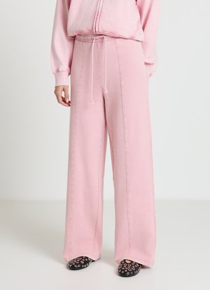 Rosa Sweatpants mit weiter Passform, Tunnelzug am Bund und sichtbaren Nahtdetails. Kombiniert mit schwarzen Slip-On-Schuhen mit Ösenakzenten.