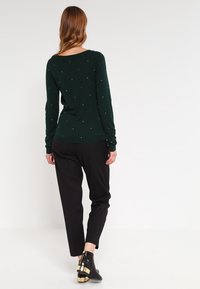 Femme aux longs cheveux portant un pull vert foncé à petits pois, un pantalon noir et des bottines noires à talons dorés, de dos.