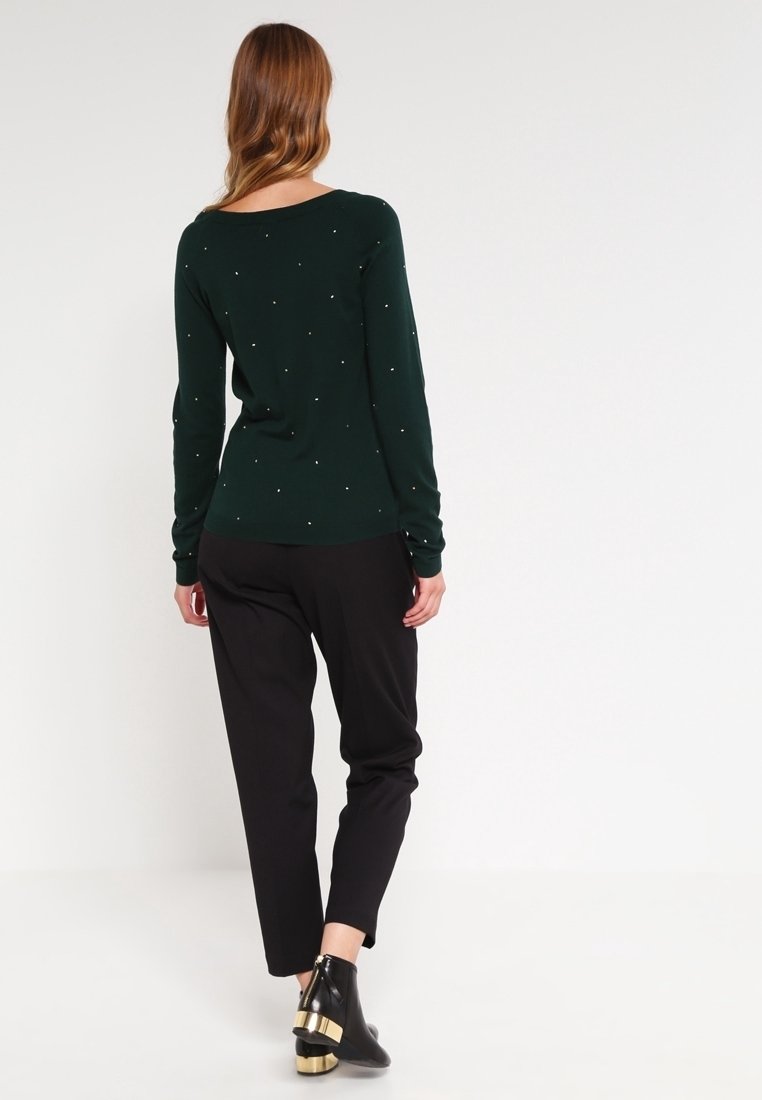 Femme aux longs cheveux portant un pull vert foncé à petits pois, un pantalon noir et des bottines noires à talons dorés, de dos.