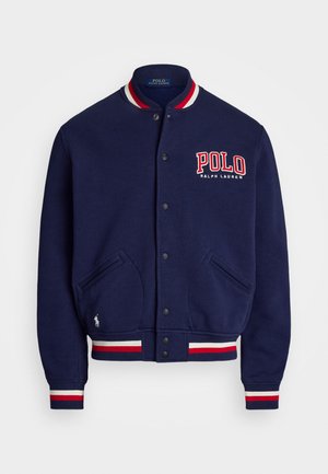 Marineblå varsityjakke med rød og hvid stribet krave og manchetter. Har en knaplukning, to frontlommer og broderet "Polo" logo.