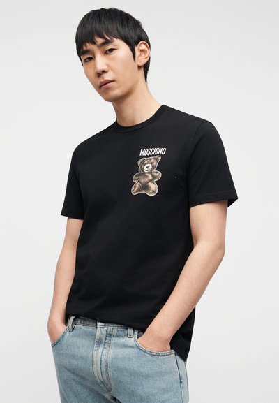 Μαύρο βαμβακερό μπλουζάκι με απεικόνιση αρκούδας στο αριστερό στήθος και το κείμενο "Moschino" από πάνω. Κλασικός στρογγυλός λαιμός και κοντά μανίκια.