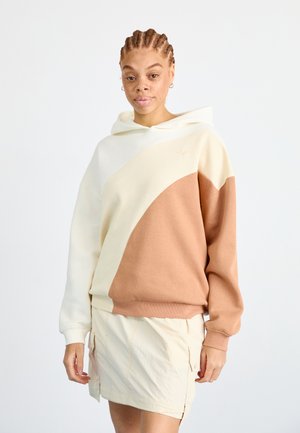 Kleurgebloekte hoodie in crème en terracotta, met een relaxte pasvorm, grote capuchon en zachte textuur. Gecombineerd met een lichtgekleurde rok.