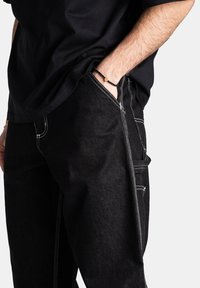 Pantaloni in denim neri con cuciture bianche a contrasto, taglio dritto e dettagli con tasche laterali. Indossati con una maglietta nera larga.