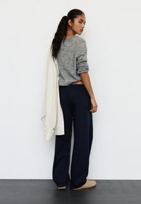 Kobieta o długich ciemnych włosach w szarym swetrze i granatowych spodniach stoi, spoglądając przez ramię, trzymając na jednym ramieniu białą koszulę w paski.