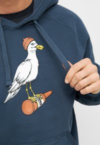 Navyblauwe hoodie met een grafisch ontwerp van een vogel met een beanie die op een fles staat, met gedetailleerde lijntekeningen en contrasterende kleuren.