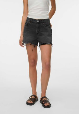 Femme portant un short en jean noir effiloché, un haut blanc sans manches et des sandales noires, debout devant un fond uni.
