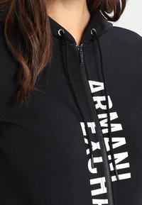 Felpa nera con zip, caratterizzata da un logo bianco ben visibile sul davanti, con lacci e un tessuto morbido e strutturato.