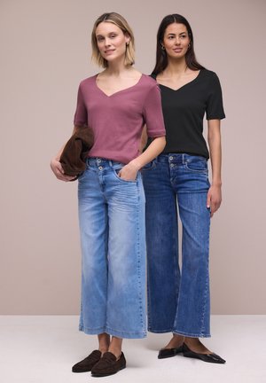 Deux femmes sont debout côte à côte, portant des hauts décontractés à col en V et des jeans en denim à jambes larges, l'une tenant une veste marron, sur un fond beige uni.