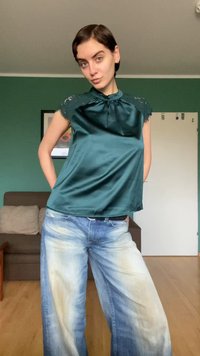 Emerald green satin blouse with lace accents at shoulders, paired with light blue, faded wide-leg jeans. Smooth texture, relaxed fit.