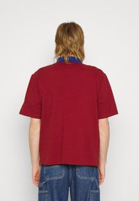 Homme aux cheveux mi-longs portant une chemise rouge à manches courtes avec un col bleu, debout de dos, portant un pantalon cargo en denim bleu visible.