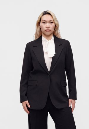 Junge Frau mit langen blonden Haaren, die eine schwarze Anzugjacke über einer weißen Bluse mit gebundener Schleife trägt und vor einem einfarbigen Hintergrund steht.