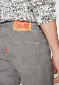 Personne portant un jean en velours côtelé gris avec une étiquette en cuir indiquant la taille et la marque, associé à une manche de pull tricot blanc et noir.