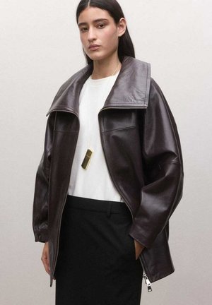 Mujer que lleva una chaqueta de cuero marrón oscuro de gran tamaño sobre una camiseta blanca y pantalones negros, combinada con un collar largo de colgante dorado.