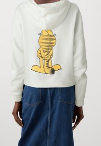 Sudadera corta blanca con un diseño de Garfield amarillos, dibujado a mano en la parte posterior. La tela es suave y tiene un corte relajado, con capucha ajustable.