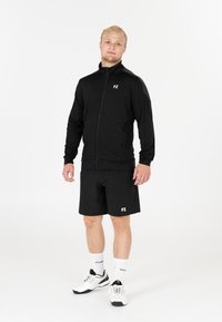 Man som står i svart zip-up jacka och shorts med vita sneakers och strumpor på en vit bakgrund.