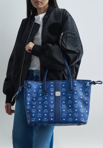 Niebieska torba na ramię z wzorzystej skóry, z logo MCM, z dwoma uchwytami i regulowanymi związami po bokach. Teksturowane wykończenie.