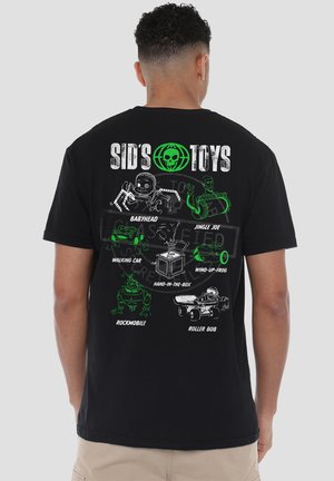 Mann trägt schwarzes T-Shirt mit grüner und weißer Sid’s Toys-Grafik mit sechs Spielzeugfiguren und Namen auf dem Rücken vor einfachem grauem Hintergrund.