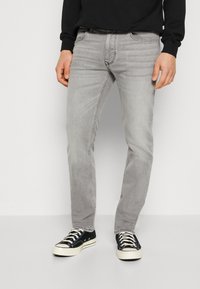 Szürke farmernadrág slim fit fazonnal, finom kopásokkal és varrás részletekkel, fekete vászon sportcipővel párosítva.