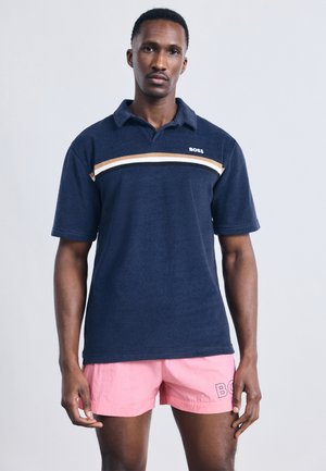 Άνδρας που φοράει μπλε σκούρο πόλο με κοντά μανίκια και ρίγες, καθώς και ροζ σορτς με λογότυπο, στέκεται μπροστά σε απλό ανοιχτό φόντο.