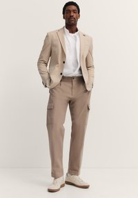 Beige maßgeschneiderter Blazer über einem weißen Hemd, kombiniert mit hellbraunen Cargo-Hosen mit Seitentaschen und cremefarbenen Sneakers mit braunen Sohlen.