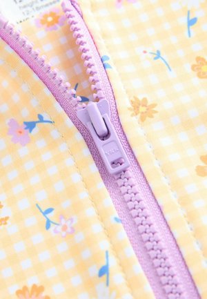 Geltonas patikrintas audinys su gėlių raštais, turintis šviesiai violetinį užtrauktuką. Lygus prigludimas su kontrastingomis siūlėmis.