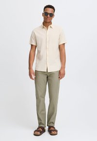 Chemise en lin beige à manches courtes, pantalon vert olive et sandales marron. Le mannequin porte des lunettes de soleil noires et se tient devant un fond uni.