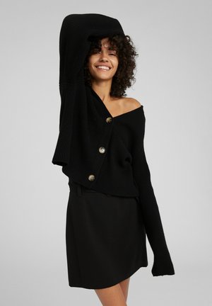 Zwarte gebreide cardigan met een off-shoulder ontwerp, voorzien van grote gouden knopen en lange mouwen. Gepaard met een zwarte rok.