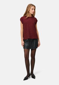 Kobieta ubrana w bordowy krótki rękaw, czarną skórzaną mini spódnicę, przezroczyste czarne rajstopy i czarne szpilki z ostrymi czubkami, stoi na białym tle.